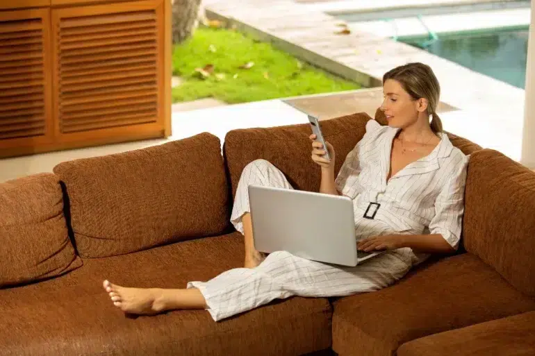 Woman in a relaxed setting, reclining on a brown sofa, using a smartphone in one hand and a laptop on her lap. She's wearing a striped outfit, with a view of a garden and a swimming pool in the background. The ambiance is calm and focused.