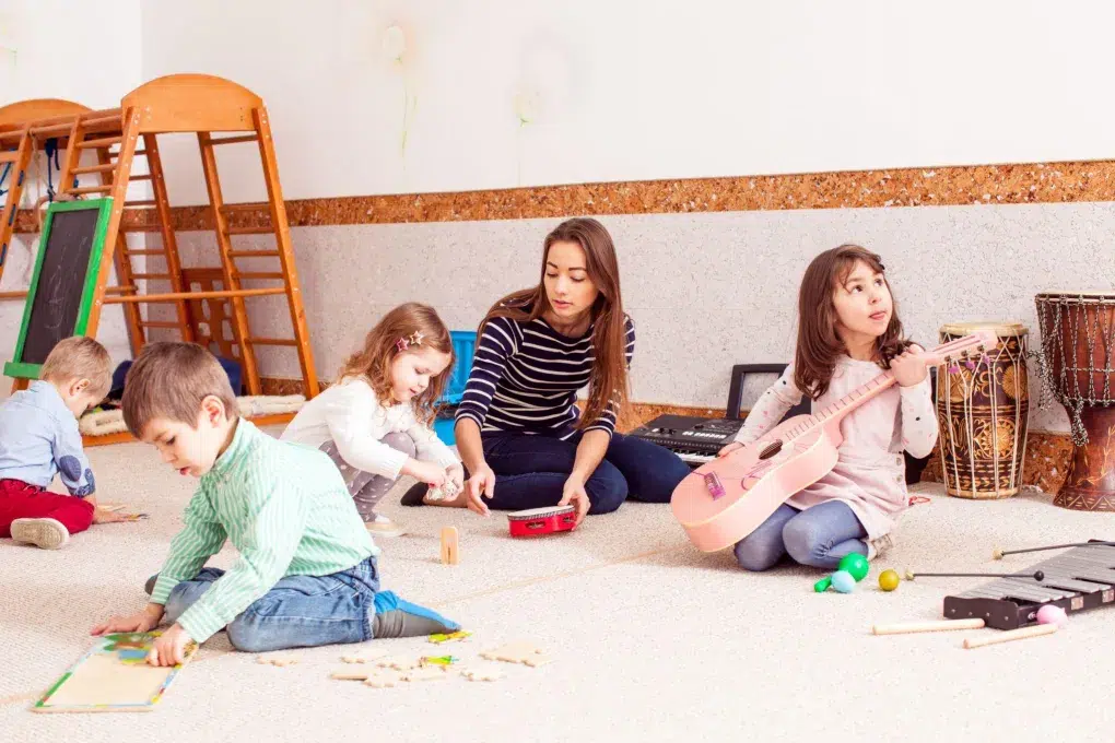 A group of children plays with toys and musical instruments on a carpeted floor in a colorful, cheerful environment.