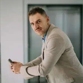 A person in a beige suit holding a smartphone, leaning slightly forward with a thoughtful expression in a modern indoor setting with neutral tones.