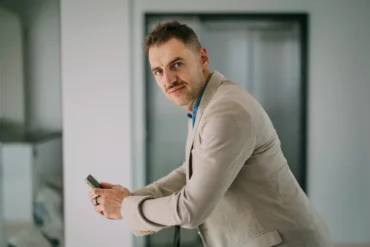 A person in a beige suit holding a smartphone, leaning slightly forward with a thoughtful expression in a modern indoor setting with neutral tones.