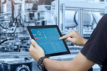 A person interacts with a tablet displaying industrial data and metrics, set against a background of machinery in a factory environment.