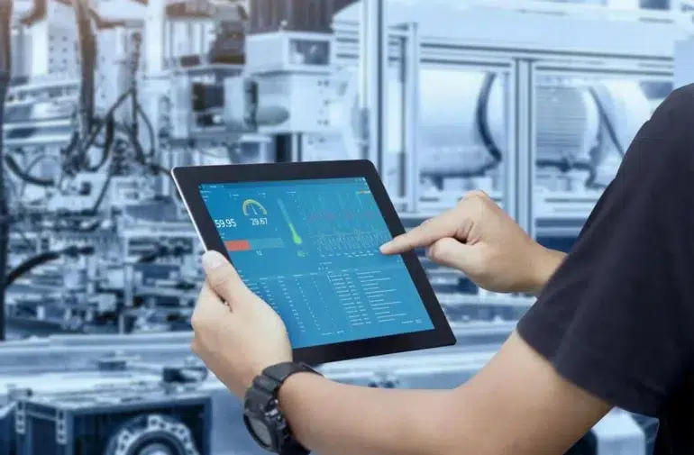 A person interacts with a tablet displaying industrial data and metrics, set against a background of machinery in a factory environment.