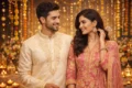 A smiling couple in elegant traditional attire stand surrounded by soft candlelight and floral decorations, creating a warm, festive ambiance.