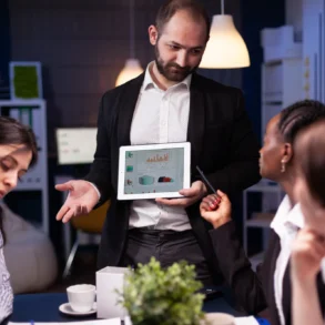 Man in a blazer presents data on a tablet to three colleagues around a table, fostering a collaborative and focused office environment.