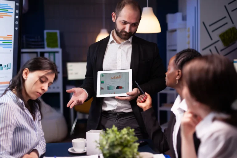 Man in a blazer presents data on a tablet to three colleagues around a table, fostering a collaborative and focused office environment.