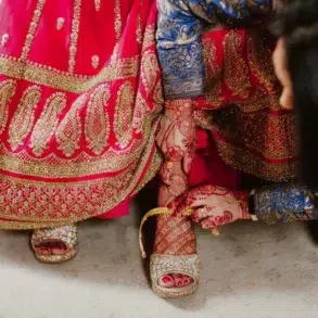 A woman in a vibrant red and gold traditional dress with intricate henna on her hands adjusts her gold high-heeled sandals, conveying elegance and cultural richness.