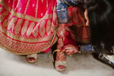 A woman in a vibrant red and gold traditional dress with intricate henna on her hands adjusts her gold high-heeled sandals, conveying elegance and cultural richness.