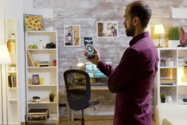 Man in a maroon shirt using a smartphone to control smart lights in a cozy, well-decorated home office.