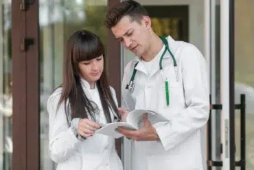 Two medical professionals in white coats review documents outside a building, with one wearing a stethoscope around the neck.