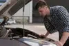 A person leans over a car engine in a parking lot, inspecting it while wearing a checkered shirt.