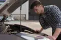 A person leans over a car engine in a parking lot, inspecting it while wearing a checkered shirt.