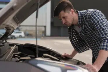 A person leans over a car engine in a parking lot, inspecting it while wearing a checkered shirt.