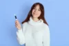 A woman in a white sweater smiles while holding an Microneedling Pen against a light blue background. The scene conveys a calm and friendly tone.