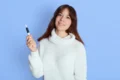 A woman in a white sweater smiles while holding an Microneedling Pen against a light blue background. The scene conveys a calm and friendly tone.