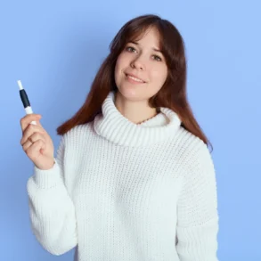A woman in a white sweater smiles while holding an Microneedling Pen against a light blue background. The scene conveys a calm and friendly tone.