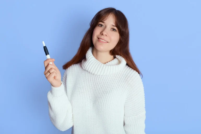 A woman in a white sweater smiles while holding an Microneedling Pen against a light blue background. The scene conveys a calm and friendly tone.
