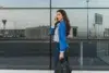 Woman in blue blazer and striped shirt talks on phone while walking, holding a black handbag. She's smiling with a glass building in the background.
