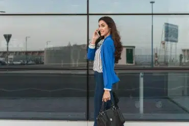 Woman in blue blazer and striped shirt talks on phone while walking, holding a black handbag. She's smiling with a glass building in the background.