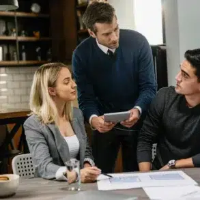 A group of three people in a modern workspace discussing plans with a tablet and documents on a table.