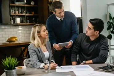 A group of three people in a modern workspace discussing plans with a tablet and documents on a table.
