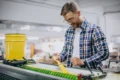 A man in a plaid shirt operates a machine in a factory, holding a yellow material. He appears focused and content. A yellow bucket is nearby.