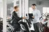 A man in a leather jacket shakes hands with a salesperson holding a clipboard in a motorcycle showroom, symbolizing a purchase agreement.