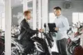 A man in a leather jacket shakes hands with a salesperson holding a clipboard in a motorcycle showroom, symbolizing a purchase agreement.