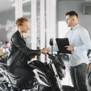 A man in a leather jacket shakes hands with a salesperson holding a clipboard in a motorcycle showroom, symbolizing a purchase agreement.