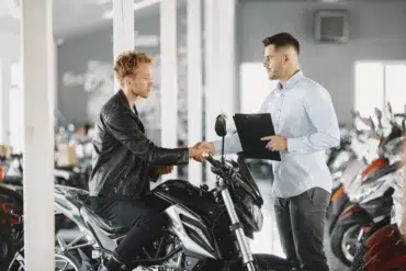 A man in a leather jacket shakes hands with a salesperson holding a clipboard in a motorcycle showroom, symbolizing a purchase agreement.