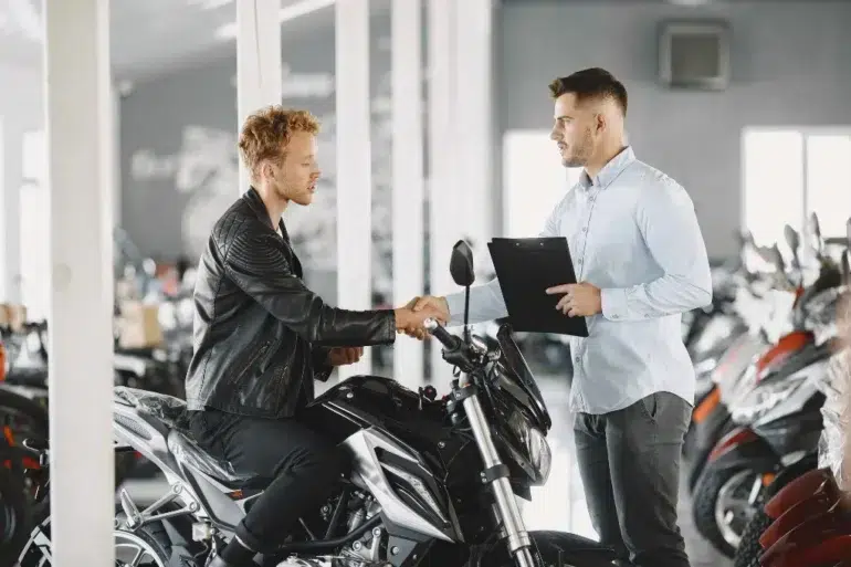 A man in a leather jacket shakes hands with a salesperson holding a clipboard in a motorcycle showroom, symbolizing a purchase agreement.