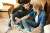 A couple sits on the floor amidst cardboard boxes, examining a house plan, with a focused expression as they discuss their new space.