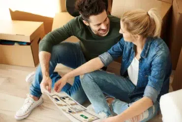 A couple sits on the floor amidst cardboard boxes, examining a house plan, with a focused expression as they discuss their new space.