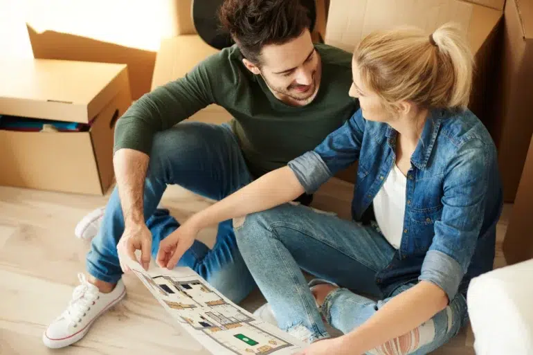 A couple sits on the floor amidst cardboard boxes, examining a house plan, with a focused expression as they discuss their new space.
