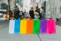 A row of colorful shopping bags in blue, yellow, orange, green, purple, and pink lined up on a busy city street.