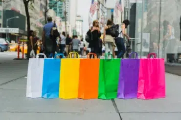 A row of colorful shopping bags in blue, yellow, orange, green, purple, and pink lined up on a busy city street.