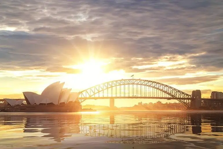 Sydney Harbour at golden hour with the Opera House and Harbour Bridge glowing in warm sunset light.