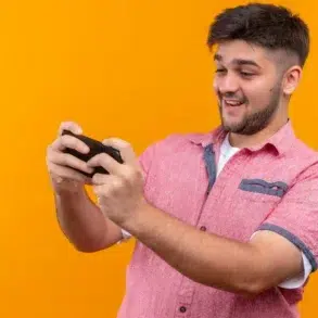 A person holding a gaming console, focused on playing, stands against a vibrant orange background.
