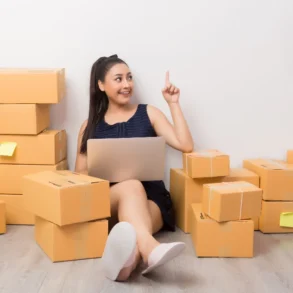 A woman sits on the floor surrounded by stacked cardboard boxes, holding a laptop. She smiles and points upward, suggesting an idea or insight.