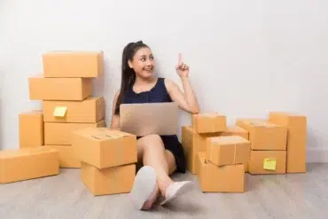 A woman sits on the floor surrounded by stacked cardboard boxes, holding a laptop. She smiles and points upward, suggesting an idea or insight.