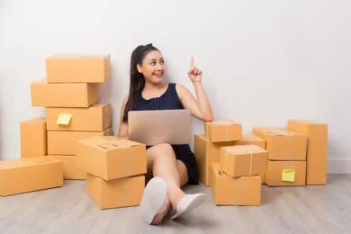 A woman sits on the floor surrounded by stacked cardboard boxes, holding a laptop. She smiles and points upward, suggesting an idea or insight.