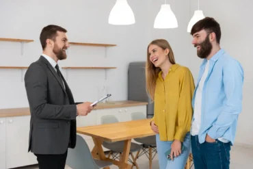 A smiling real estate agent with a clipboard talks to a happy couple in a modern kitchen. The room has a wooden table, shelves, and pendant lights.