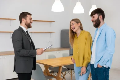 A smiling real estate agent with a clipboard talks to a happy couple in a modern kitchen. The room has a wooden table, shelves, and pendant lights.