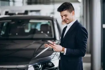 A man in a pinstriped suit stands near a black car, holding a tablet and engaged in a conversation.