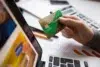 A hand holds a green and gold credit card above a laptop keyboard, surrounded by office supplies and documents.