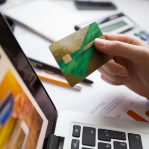 A hand holds a green and gold credit card above a laptop keyboard, surrounded by office supplies and documents.