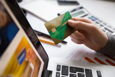 A hand holds a green and gold credit card above a laptop keyboard, surrounded by office supplies and documents.
