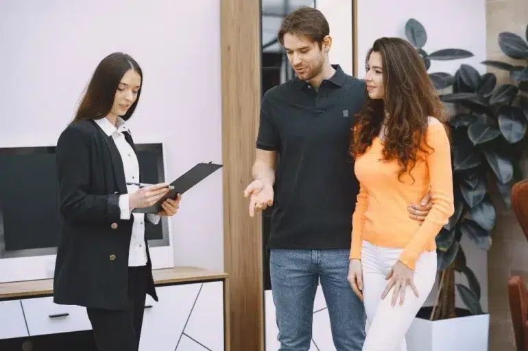 A couple stands closely while a professional woman holds a clipboard and explains something in a modern office setting.