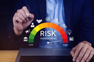 A person in a suit holds a pen above a risk management gauge, ranging from low to high, with business icons. The tone is professional and analytical.