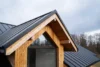Detail of a modern house roof featuring a wooden gable and dark metal shingles against a cloudy sky.