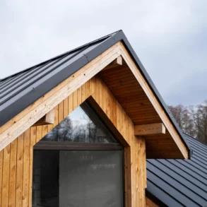 Detail of a modern house roof featuring a wooden gable and dark metal shingles against a cloudy sky.
